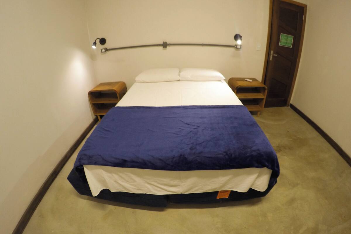Ett standardhotellrum med en säng, två sängbord, ett litet bord, en vägglampa och en dörr med en skylt.