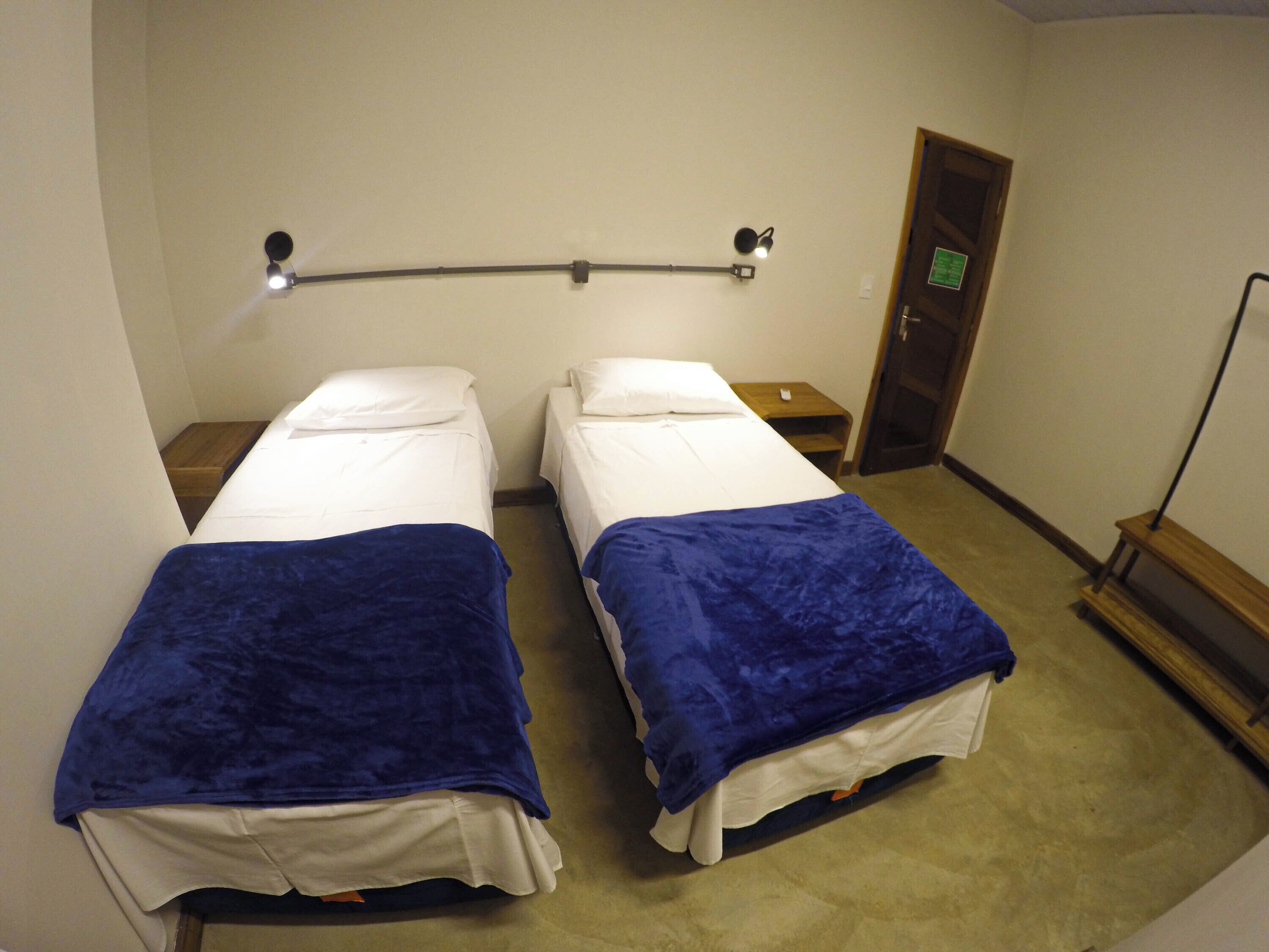 Una habitación con dos camas, cada una con una manta azul y sábanas blancas, una mesita de noche con lámpara y una puerta al fondo.