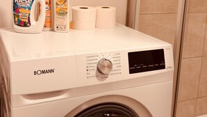 Un lave-linge à chargement frontal, doté d’un panneau de commande, avec une bouteille de lessive, une bouteille d’adoucissant et deux rouleaux de papier toilette posés sur le dessus.