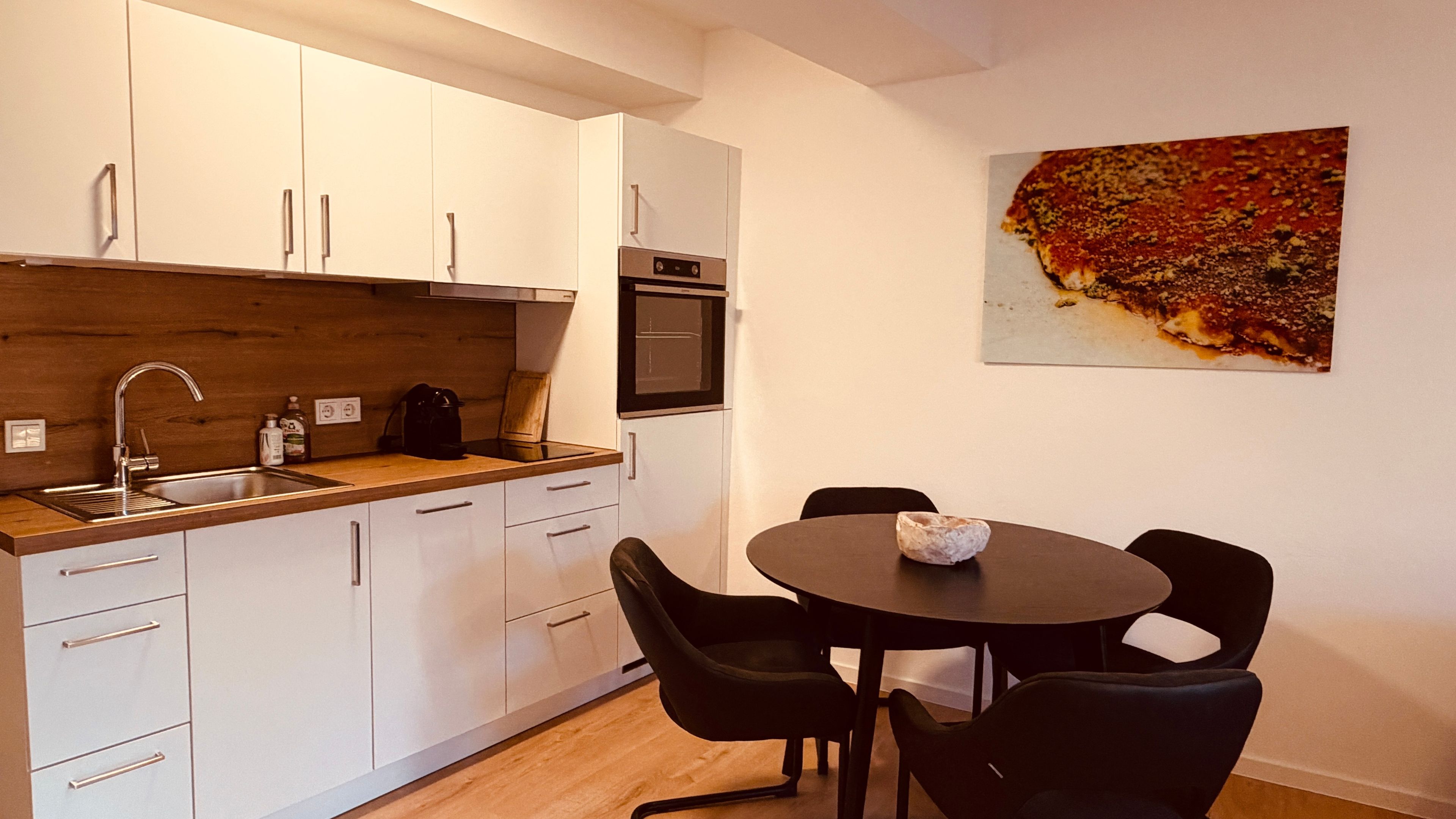 Eine kleine Küche mit weißen Schränken, einer Holz Arbeitsplatte, einem Spülbecken, einer Kaffeemaschine und einem kleinen Backofen. Ein Esstisch mit vier Stühlen und einem einzelnen Dekorationselement steht daneben. Ein Gemälde hängt an der Wand über dem Tisch.
