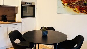 Une cuisine compacte avec des meubles blancs, un plan de travail noir et un four encastré. On y trouve une table ronde avec quatre chaises noires et un petit vase bleu. Un grand tableau abstrait est accroché au mur.