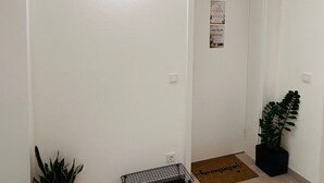 A hallway with a white wall, a door, a potted plant, a black wire shelf, and a doormat. - Cozy Corner – Passau (Passau)