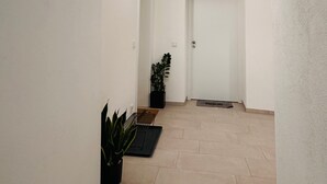 Un couloir avec une porte blanche au fond, une plante en pot et un luminaire fixé au mur.