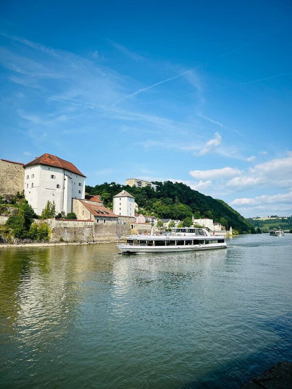 A river with a boat, buildings along the riverbank, and a hill in the background. - The Cozy Corner – Passau (Passau)