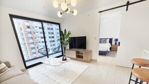 A modern living room with a large sliding glass door, a flat-screen TV, a wooden TV stand, a potted plant, a white rug, a sofa, and a bar stool. - Modern and cozy apartment in the heart of Panama City, ideal for business travelers and digital nomads. Pool, gym, and coworking space available, with valet parking offered as an optional service. (Panamá)