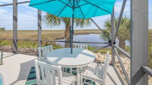A covered outdoor seating area with a turquoise umbrella, white chairs, and a view of a water body and palm trees.