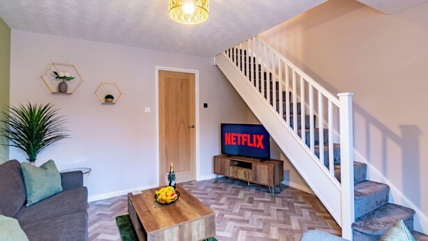 A living room with a sofa, coffee table, and a TV displaying 'NETFLIX'. A staircase with white railings leads to another floor.
