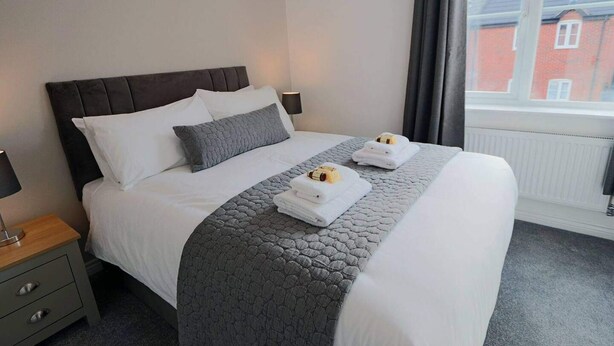 Una cama doble con arreglos ordenados, ropa de cama blanca y gris, dos toallas sobre la cama, un edredón gris y una ventana con vista a un edificio rojo.