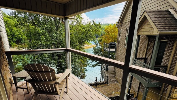 Une terrasse en bois offrant une vue sur une rivière et des arbres, un espace couvert avec une chaise et une table, et un petit bâtiment doté d’un toit en pente.