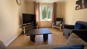 A living room with a flat-screen TV, a coffee table, two sofas, a chair, and a window with curtains.