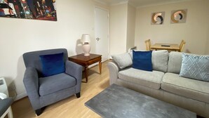 A living room with a grey armchair, a grey sofa with blue cushions, a small wooden side table with a lamp, and a grey coffee table.