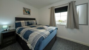 Un dormitorio con cama, mesita de noche, lámpara, ventana con cortinas y una obra de arte en la pared.