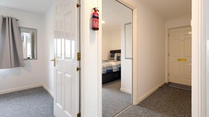 Un pasillo con una puerta blanca, un extintor y una cama en la habitación contigua.
