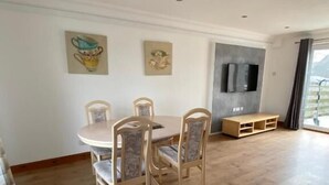 A dining area with a round table and four chairs, a flat-screen TV mounted on the wall, and a balcony with a view.