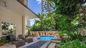 Uma área à beira da piscina com mesa e cadeiras, cercada por plantas em vasos e um edifício alto ao fundo.
