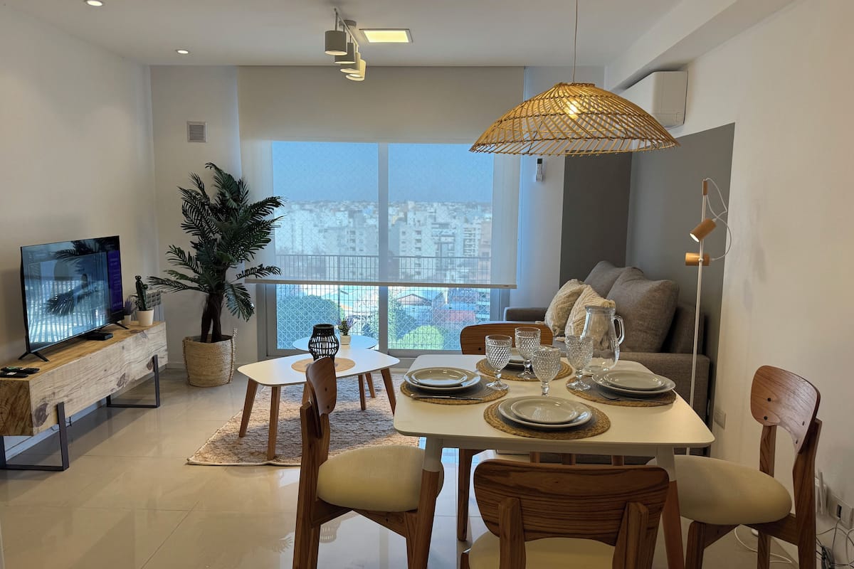 A modern dining area with a large window, a dining table set with plates and glasses, and a television on a wooden console.