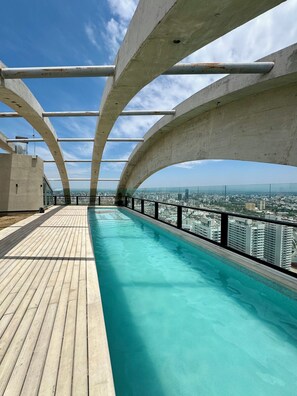 A rooftop swimming pool with a city view, surrounded by a wooden deck and metal railings.