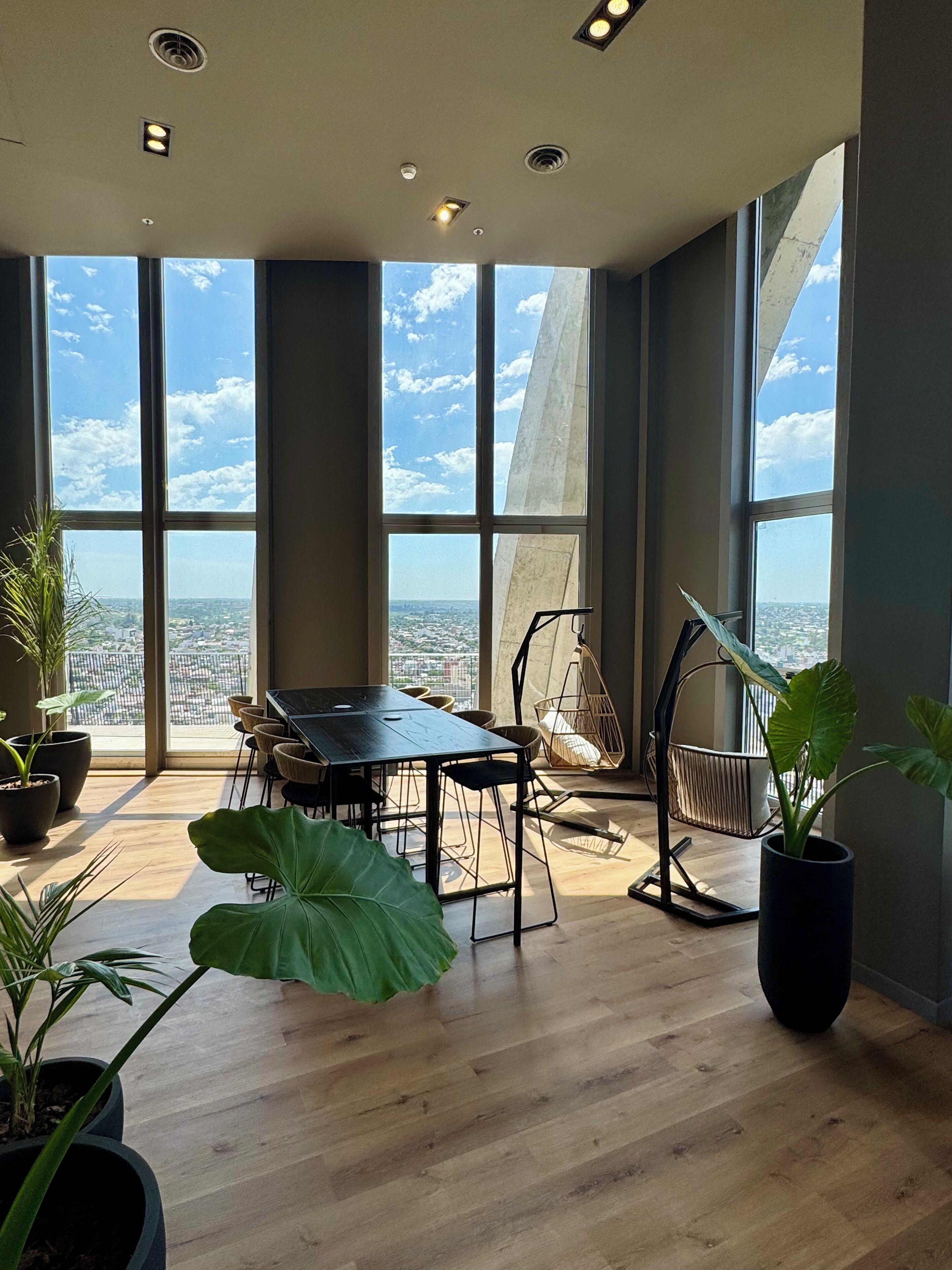 A spacious room with a large table and chairs, large windows with a city view, and several potted plants.