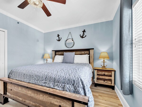A bedroom with a bed, wooden headboard, two bedside tables with lamps, a ceiling fan, and wall decorations.