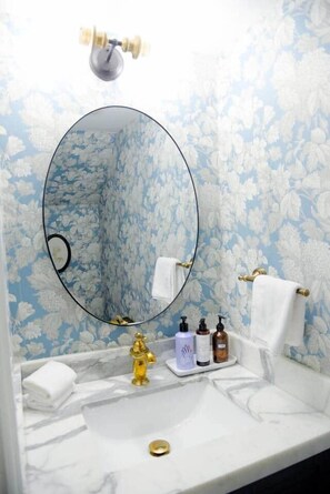 A bathroom with a marble sink, gold faucet, and a round mirror. There are toiletries on the counter and a towel rack with white towels.