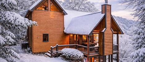 A log cabin with a porch and a chimney, surrounded by snow-covered trees.
