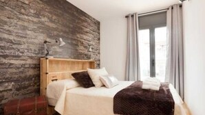 Une chambre à coucher avec une tête de lit en bois, un lit recouvert d’une literie blanche, un plaid de couleur marron foncé, une lampe et une valise.