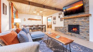 A cozy living room with a fireplace, a television mounted on the wall, and a dining area in the background.