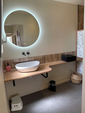 A modern bathroom with a white sink, a round mirror with a light, a wooden countertop, and a towel rack.