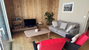 A cozy living room with a wooden wall, a grey sofa, a red chair, a wooden coffee table, and a television. - Gîte Ondine - House with indoor pool (Belz)