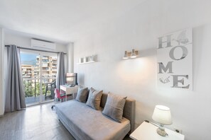 A modern hotel room with a large bed, a desk, a chair, a lamp, and a balcony with a view of buildings.