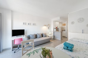 A modern hotel room with a flat-screen TV, a sofa, a dining table, and a bed with rolled towels.