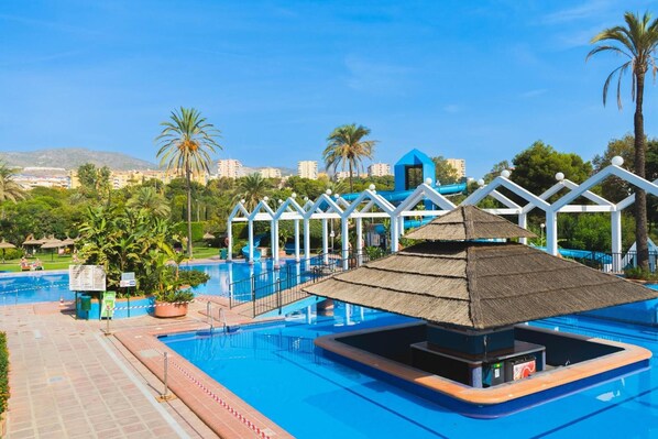 A swimming pool with a thatched-roof structure, surrounded by palm trees and a clear blue sky.