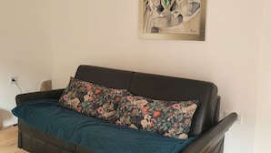 Ein Wohnzimmer mit einem schwarzen Ledersofa, einem Blumenkissen und einer blauen Decke. Darüber an der Wand hängt ein Gemälde.