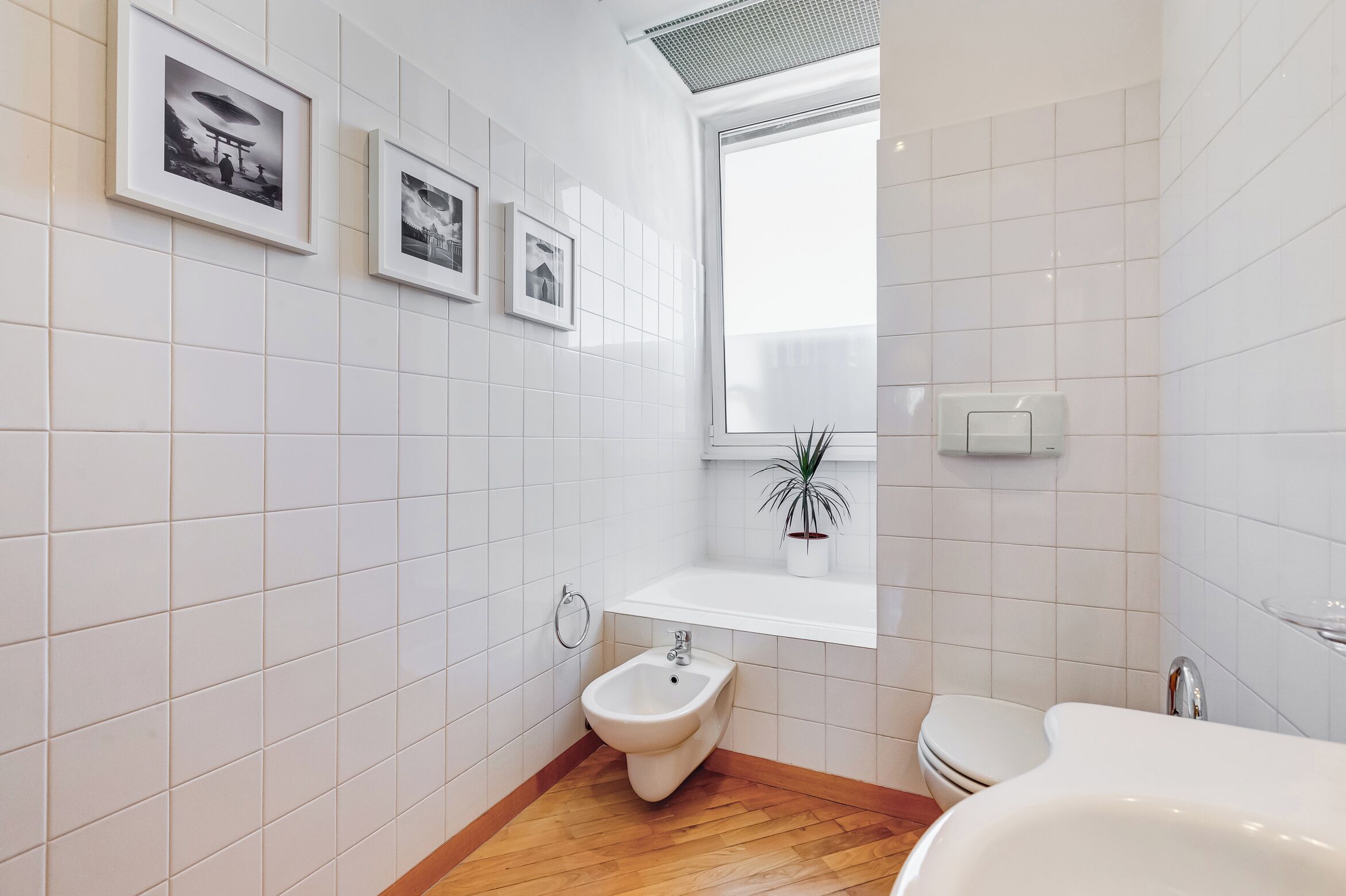Een moderne badkamer met witte tegels, een bidet, een toilet en ingelijste foto’s aan de muur.
