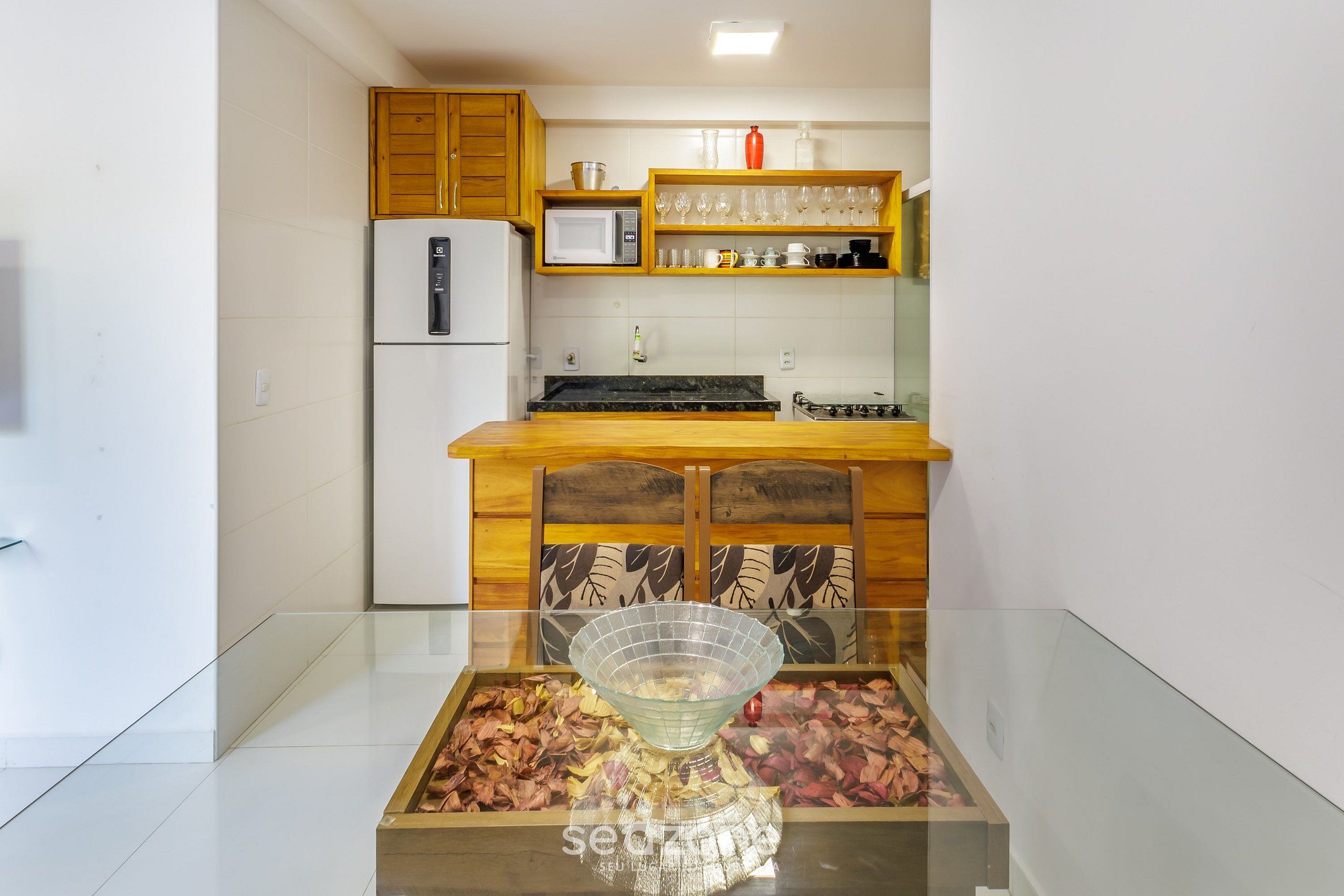 A kitchen with a refrigerator, microwave, wooden cabinets, and a glass table with a decorative bowl.