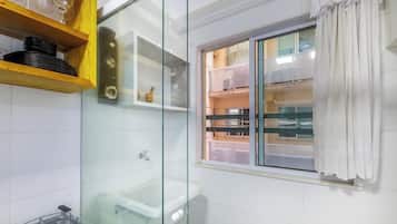 Un baño con cabina de ducha de vidrio, inodoro y ventana con vistas a un edificio.