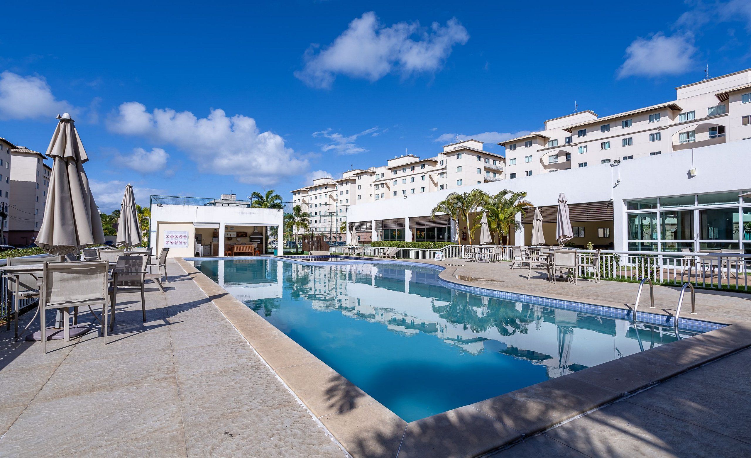 A swimming pool with lounge chairs and umbrellas, surrounded by buildings with balconies and a clear sky.