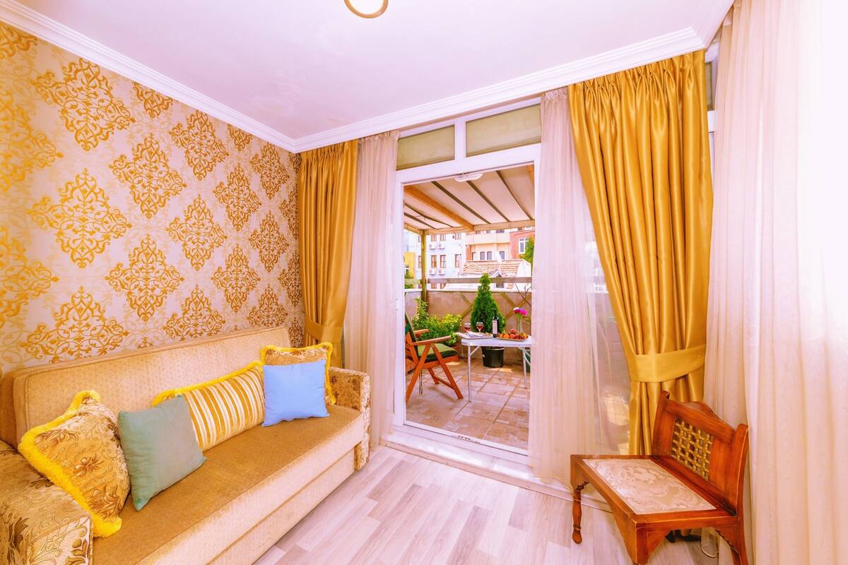 A room with a patterned wall, a sofa with cushions, a wooden chair, and a view through a glass door to an outdoor area with plants and a table.