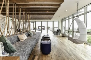 Un vaste espace de vie doté d’un plafond en bois, d’un fauteuil suspendu, d’un canapé avec des coussins et d’une grande fenêtre offrant une vue sur la ville.