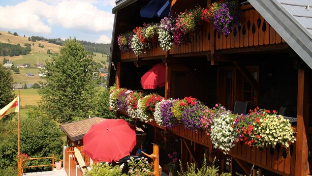 A wooden building with a balcony adorned with flower baskets and a red umbrella. - Panorama Apartment 2 – NEW! incl. 3 Worlds Card – 140 Free Attractions (Bernau im Schwarzwald)