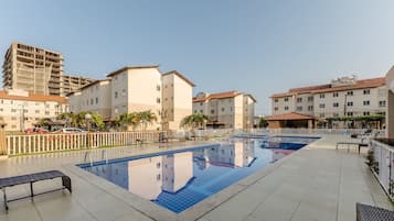 A swimming pool with a tiled deck, surrounded by buildings with balconies and a clear sky.