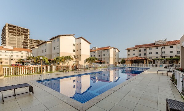 A swimming pool with a tiled deck, surrounded by buildings with balconies and a clear sky.