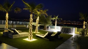 Un área de descanso en la azotea con palmeras iluminadas, sillones y vistas a la ciudad de noche.