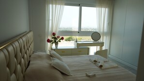 Una cama bien hecha con almohadas y una toalla doblada, una mesita de noche con espejo, un jarrón con flores y vista a la ciudad por la ventana.