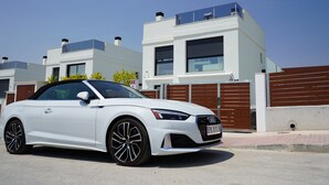 Un Audi convertible blanco estacionado frente a una casa moderna de dos pisos, con un camino de ladrillos.