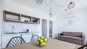 A modern kitchen with white cabinets, a dining table with a bowl of fruit, and a grey sofa in the living area.