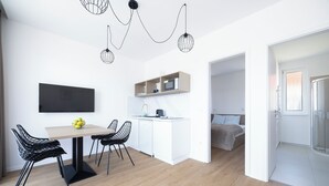 A modern kitchen with a microwave, a coffee machine, and a small shelf. There is a dining table with chairs and a bowl of fruit. The room has wooden flooring and white walls.