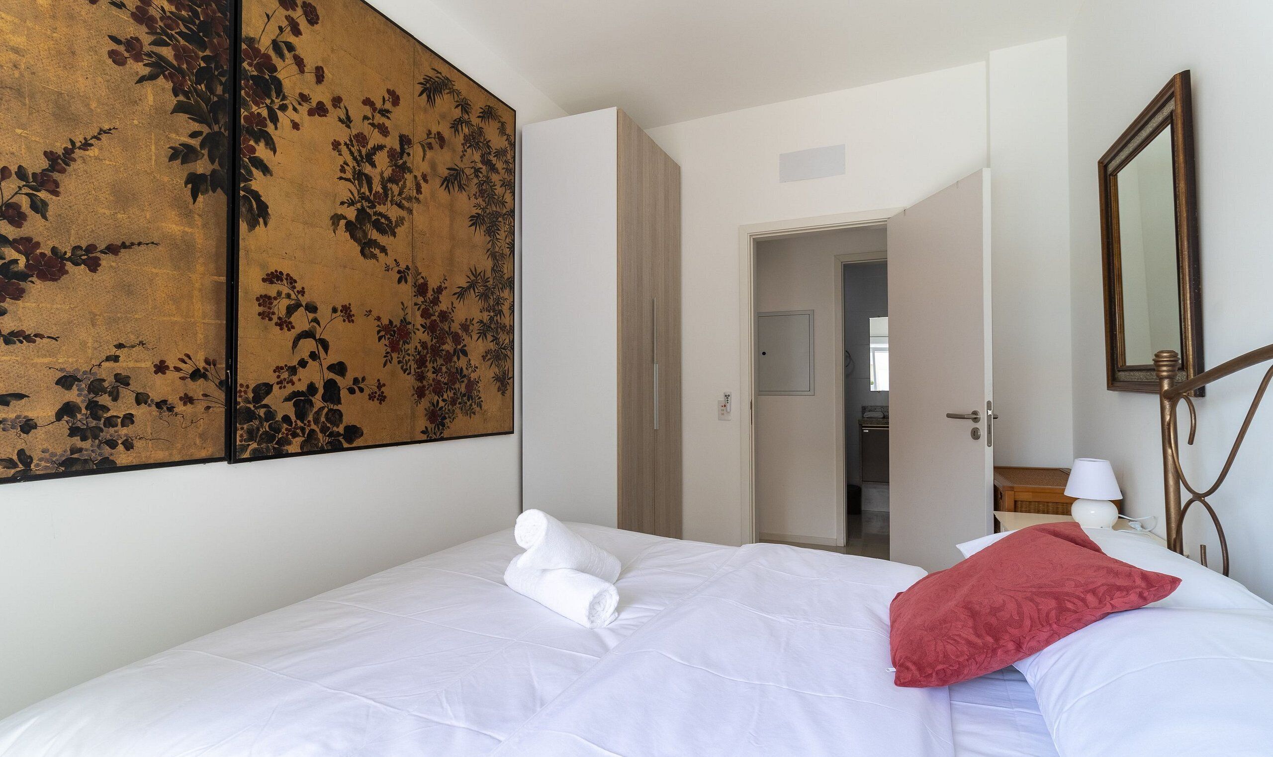 Hotellihuone, jossa on sänky, suuri kukka-aiheinen taideteos seinällä, peili ja ovi, joka johtaa toiseen huoneeseen.