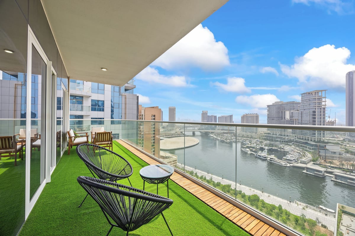 Een balkon met kunstgras, zwarte wickerstoelen en een kleine tafel. Achtergrond toont een rivier met boten en gebouwen.