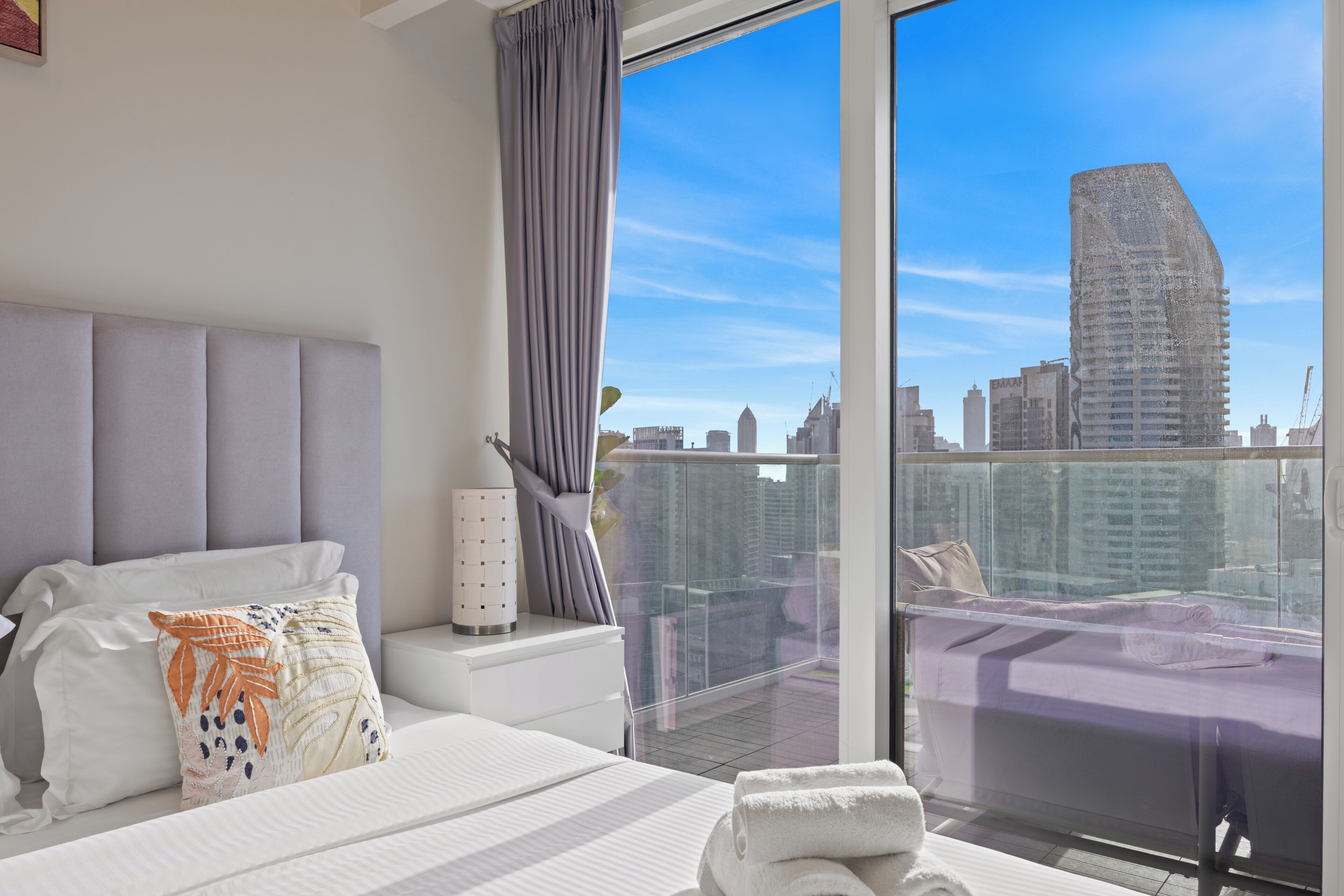 A modern hotel room with a large bed, a bedside table, a nightstand, a window with a city view, and a balcony with a view of the city skyline.